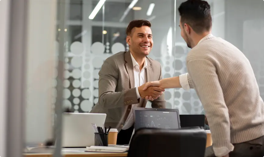 Two professionals shaking hands over a desk in a modern office.