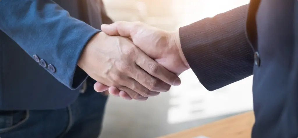 Two individuals shaking hands over a contract on a desk.